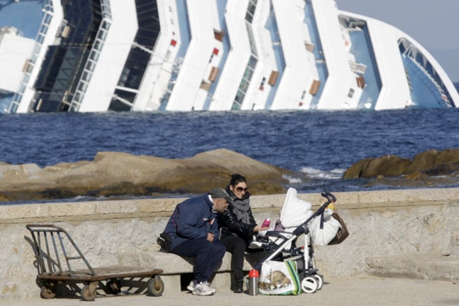 A couple feeds their child while sitting on a dock in front of the Costa Concordia cruise ship that 