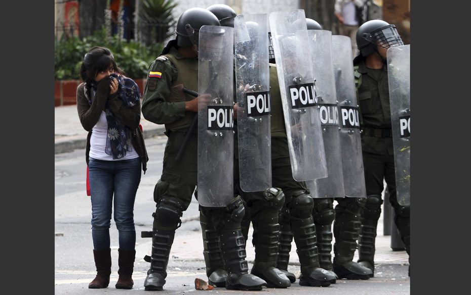 A woman takes cover behind riot police as protesters throw stones during a demonstration against wha