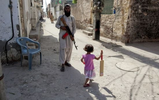 A little girl with a cricket bat walks past a guard of Malik Ishaq, the leader of Lashkar-e-Jhangvi 