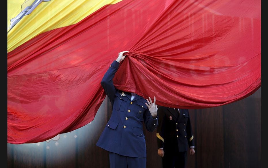 A member of the Spanish Armed Forces gets his face covered by a giant Spanish flag as he helps raise