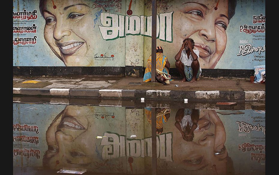 A couple sits along a flooded roadside under a picture of Tamil Nadu Chief Minister Jayalalithaa in 