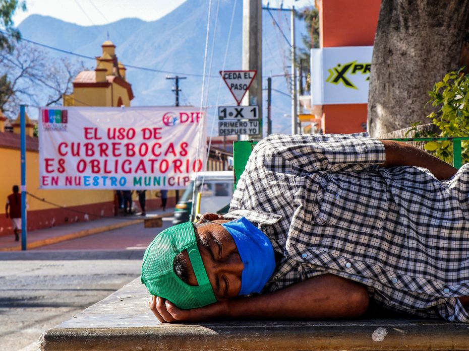 A man wearing a face mask sleeps at a park near a banner reading 