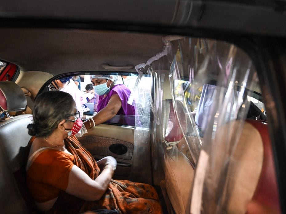 A health worker stretches herself to vaccinate an elderly woman through the car’s window at a 