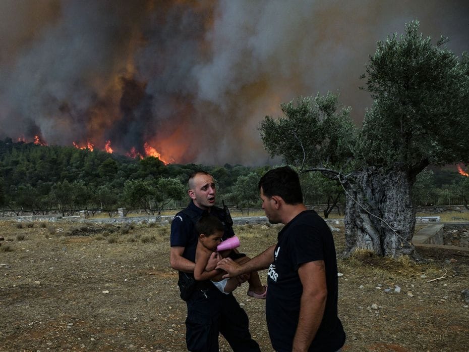 A police officer evacuates a child from a house amidst ancient olive trees as a wildfire burns, in t